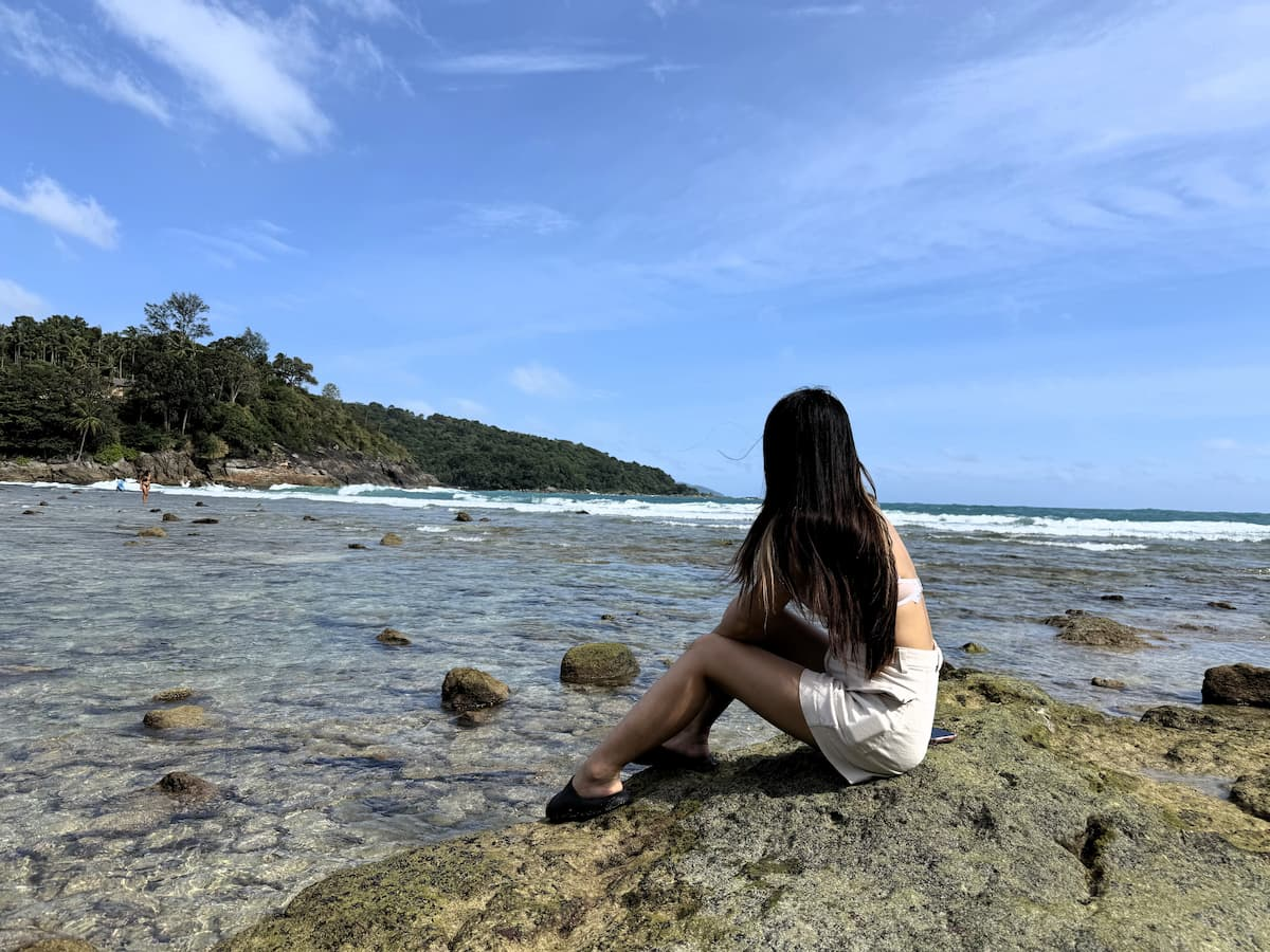 Visitor sitting on the rocky shoreline at Merlin Beach with the tropical headland in the background