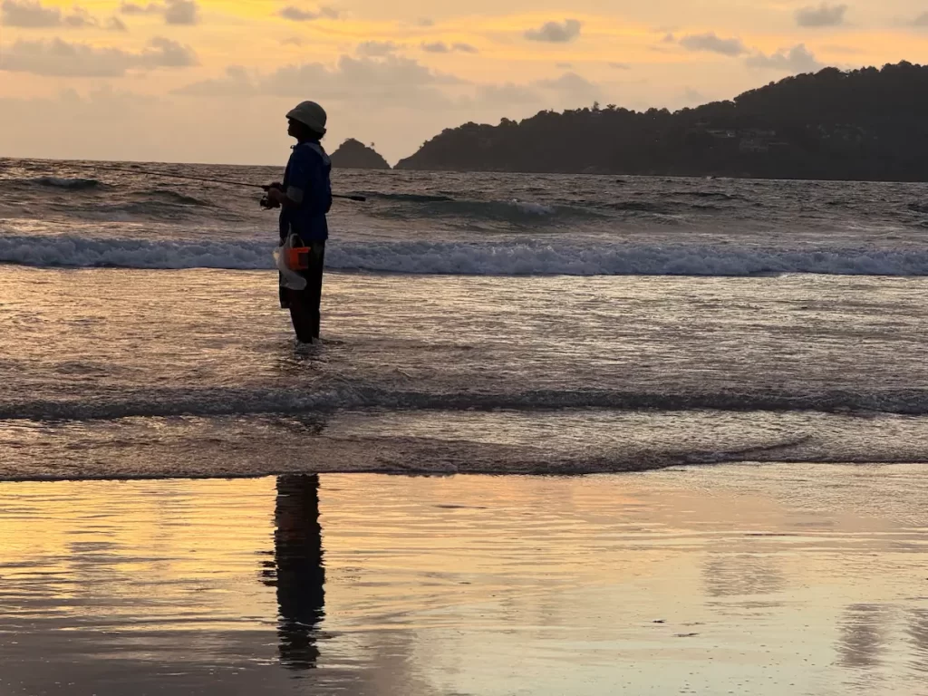 A fisherman standing in the shallow water at Patong Beach during sunset, with the Andaman Sea and headland glowing in the evening light.