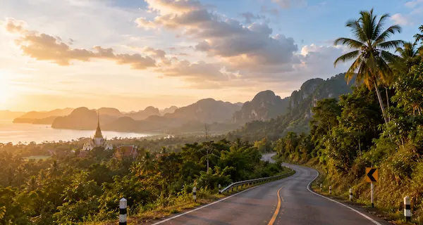 Thailand travel guide showing a scenic road winding through tropical landscapes towards a traditional Thai temple and dramatic coastline.