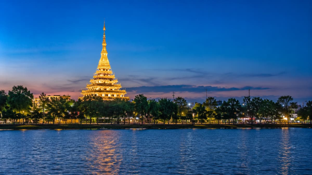 Thailand travel guide view across Bueng Kaen Nakhon towards the illuminated Wat Nong Wang and Khon Kaen waterfront at blue hour.