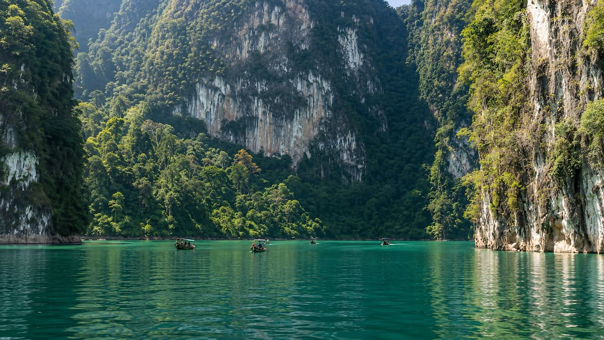 Thailand travel guide view of long-tail boats crossing Cheow Lan Lake beneath rainforest-covered limestone cliffs in Khao Sok.
