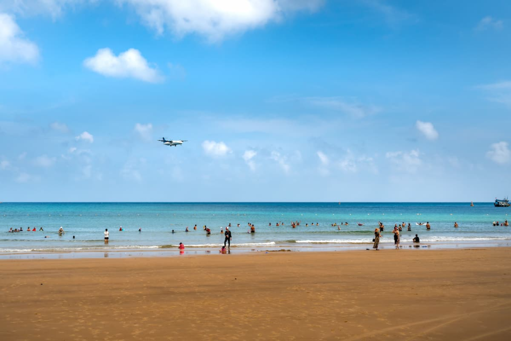 Families swimming at Mai Khao Beach as an aircraft approaches Phuket International Airport.