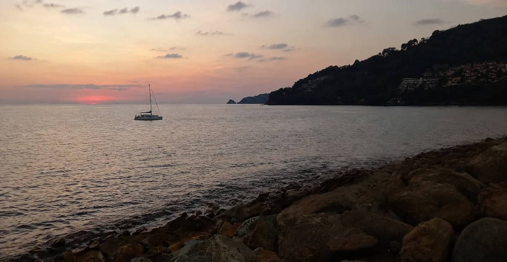 Anchored catamaran at sunset off Kalim Beach near Patong, Phuket, with a rocky shoreline and forested headland.
