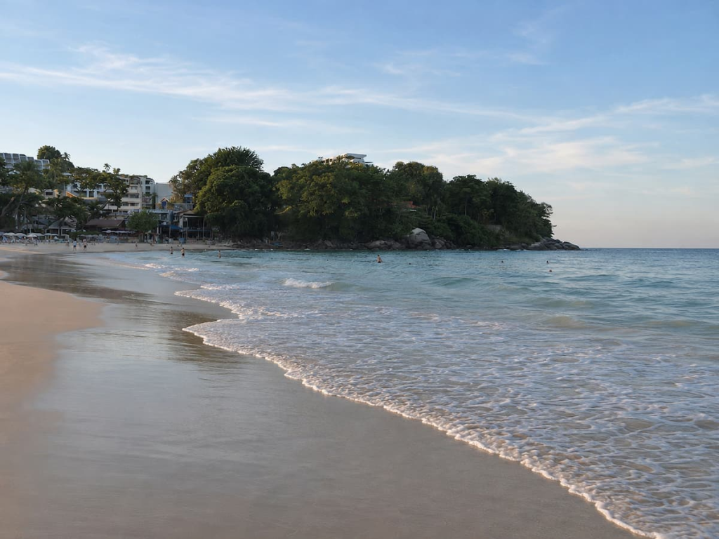 Waves washing onto Kata Beach beside a wooded headland and beachfront buildings, in our Phuket travel guide.
