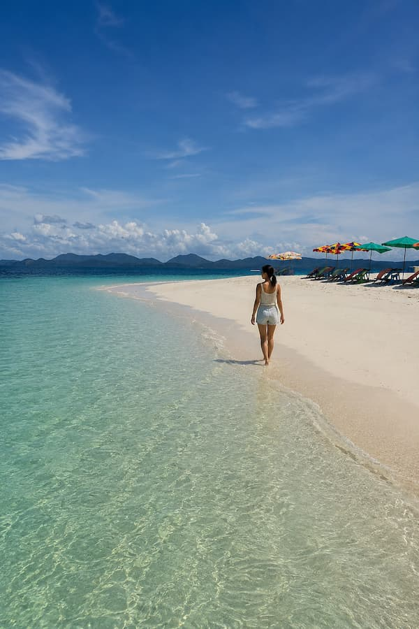 Woman walking beside clear shallow water and white sand in the Khai Islands, featured in our Phuket travel guide.