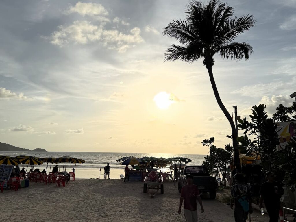 Patong Travel Guide view of the beachfront at sunset, with palm trees, umbrellas, vendors and water-sports activity on Patong Beach.