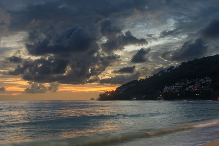 Patong weather changing at sunset, with dark tropical clouds over the bay, waves reaching the beach and the southern headland in view.
