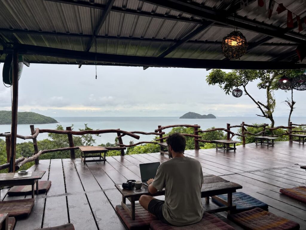 Traveller using a laptop to plan a multi-destination Thailand holiday from a quiet coastal viewpoint.