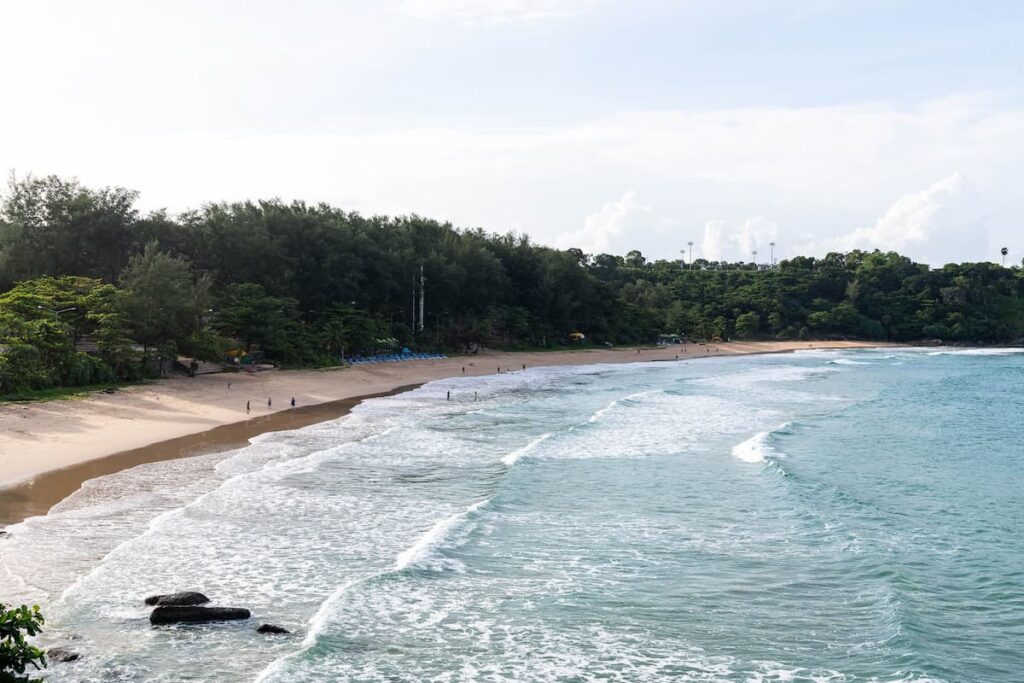 Sandy beach with turquoise water and forested coastline in Phuket, Thailand.